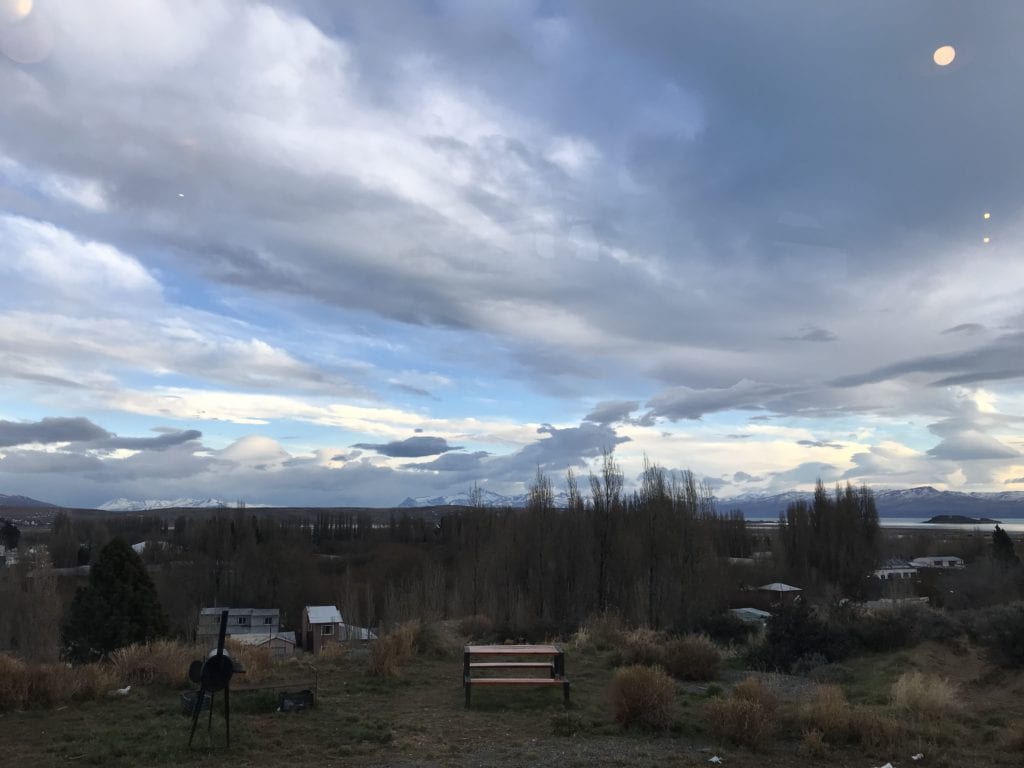 view of a landscape, bench and grill in the foreground; leafless trees in the midground; mountains and blue sky with majestic clouds in the background